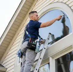 Professional window cleaner on a ladder using a squeegee to clean an exterior arched window, Columbus, OH.